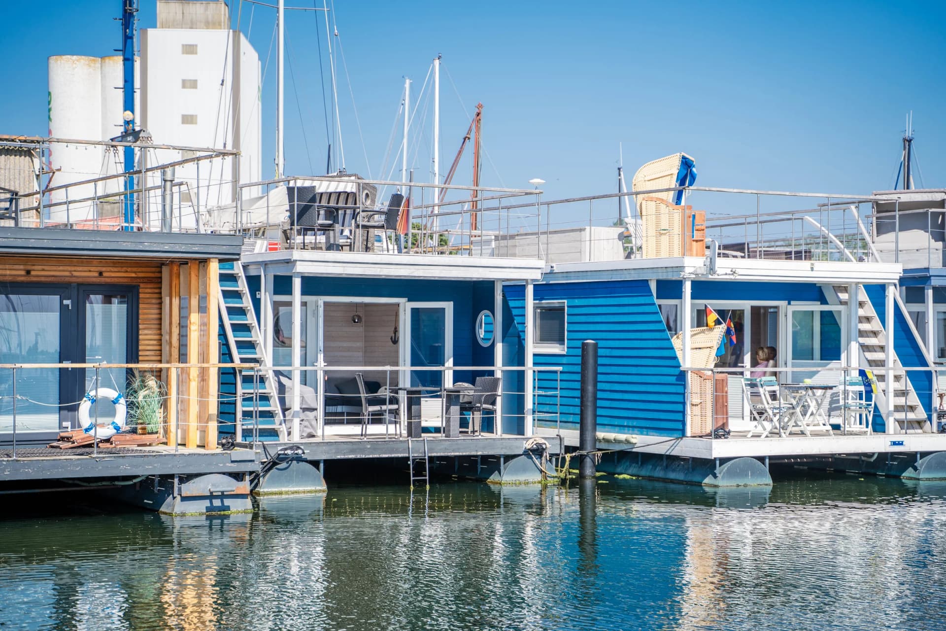 Blau-weiße Hausboote im Ostseehafen, daneben Segelboote und Hafenkulisse